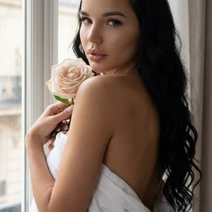 Ultra-realistic iPhone portrait near a bright window in an elegant Parisian apartment. The woman sits wrapped in a white sheet with her back turned slightly while looking over her shoulder toward the camera. She holds a pale blush rose near her shoulder. 