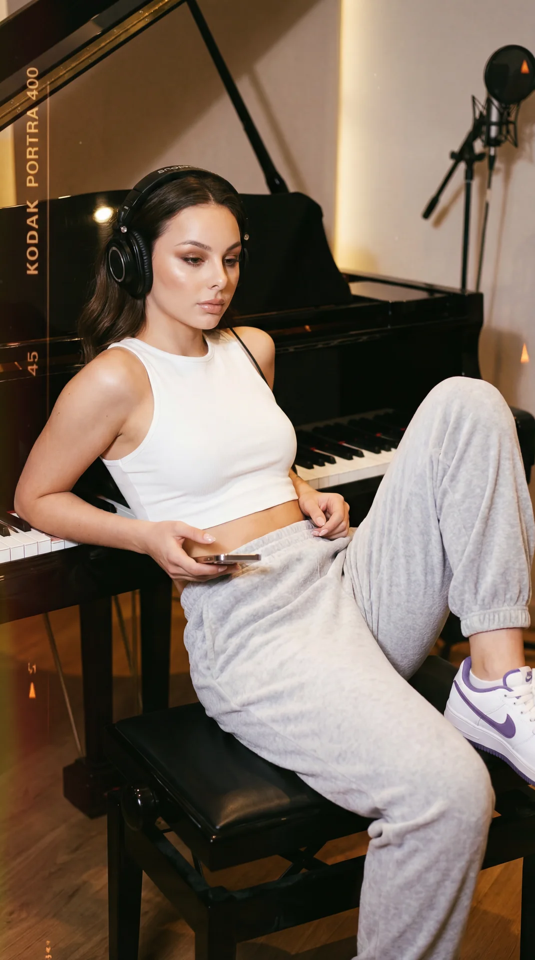 A girl is captured in a relaxed, candid medium shot leaning back on a black piano bench in a recording studio. The camera angle is slightly high and tilted, emphasizing her casual, comfortable pose and the environment around her. She reclines on the bench