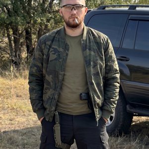 Высококачественное фото молодого мужчины в стиле military. На нем объемная темно-зеленая камуфлированная куртка-бомбер, надетая поверх оливковой базовой футболки, и широкие черные тактические штаны с карманами, на правом бедре тактический нож в ножнах. Из