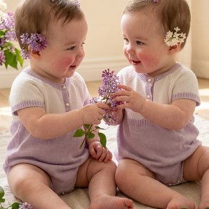 Ultra-realistic professional photo of two identical twin baby girls, 8 months old. They are sitting on a soft cream linen rug, wearing matching beautiful knitted bodysuit dresses in pale lilac and white with tiny pearl buttons. Fresh lilac flowers (syring