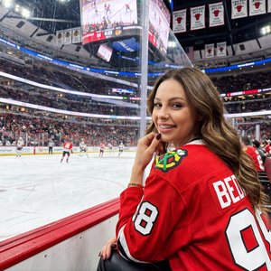 Create a realistic high quality photo using the face from the reference without any changes or distortions. Realistic lifestyle photo of a young woman at a hockey game in a huge modern arena. The atmosphere is lively energetic and flirty with American spo