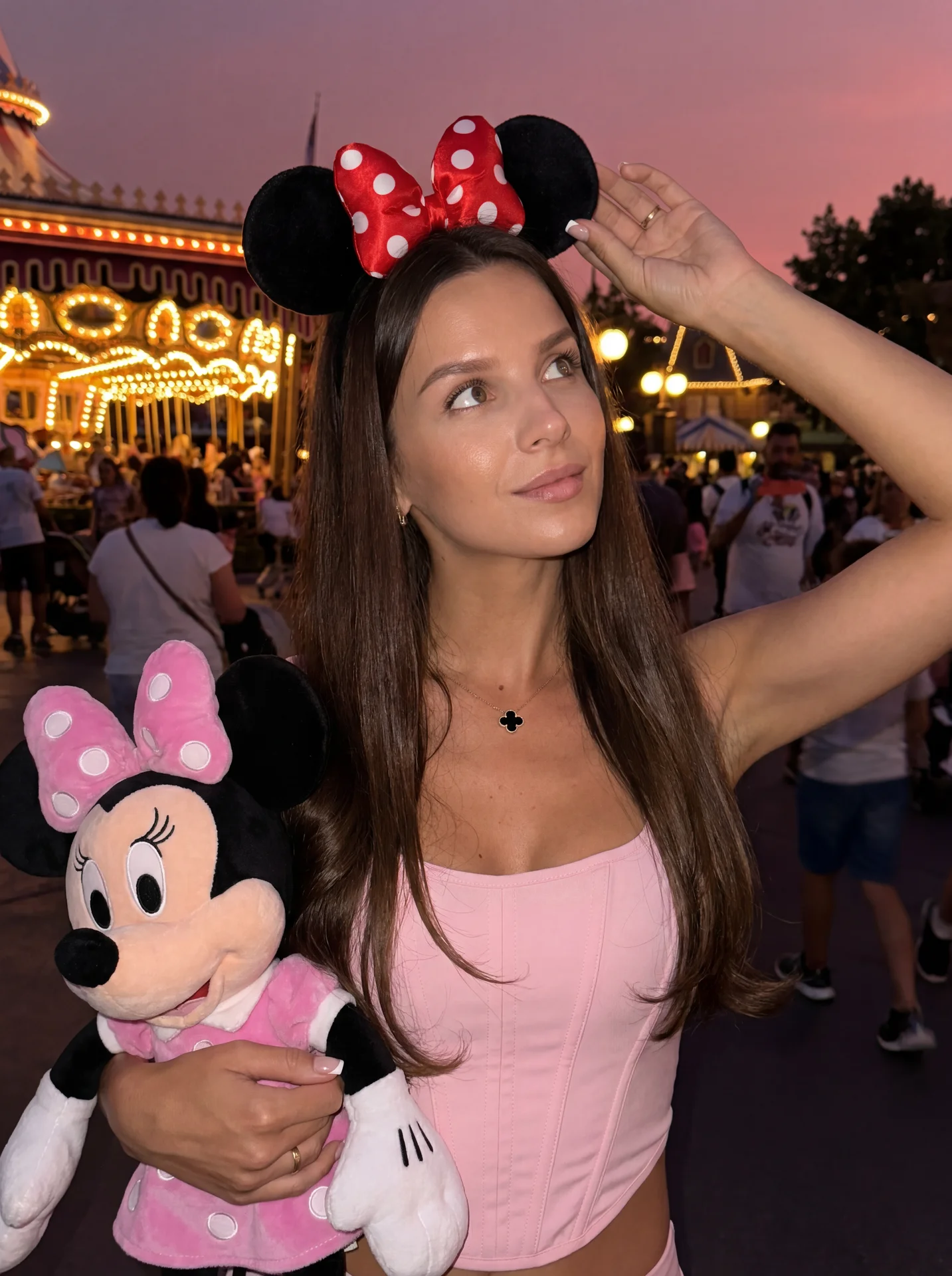 Не меняя внешности девушки с фото, девушка стоит вечером в парке Disney, держит в руке большую игрушку Minnie Mouse в розовом платье.
на голове ободок Minnie Mouse с красным бантом в белый горошек и черными ушками.
она смотрит вверх в сторону, одной рукой
