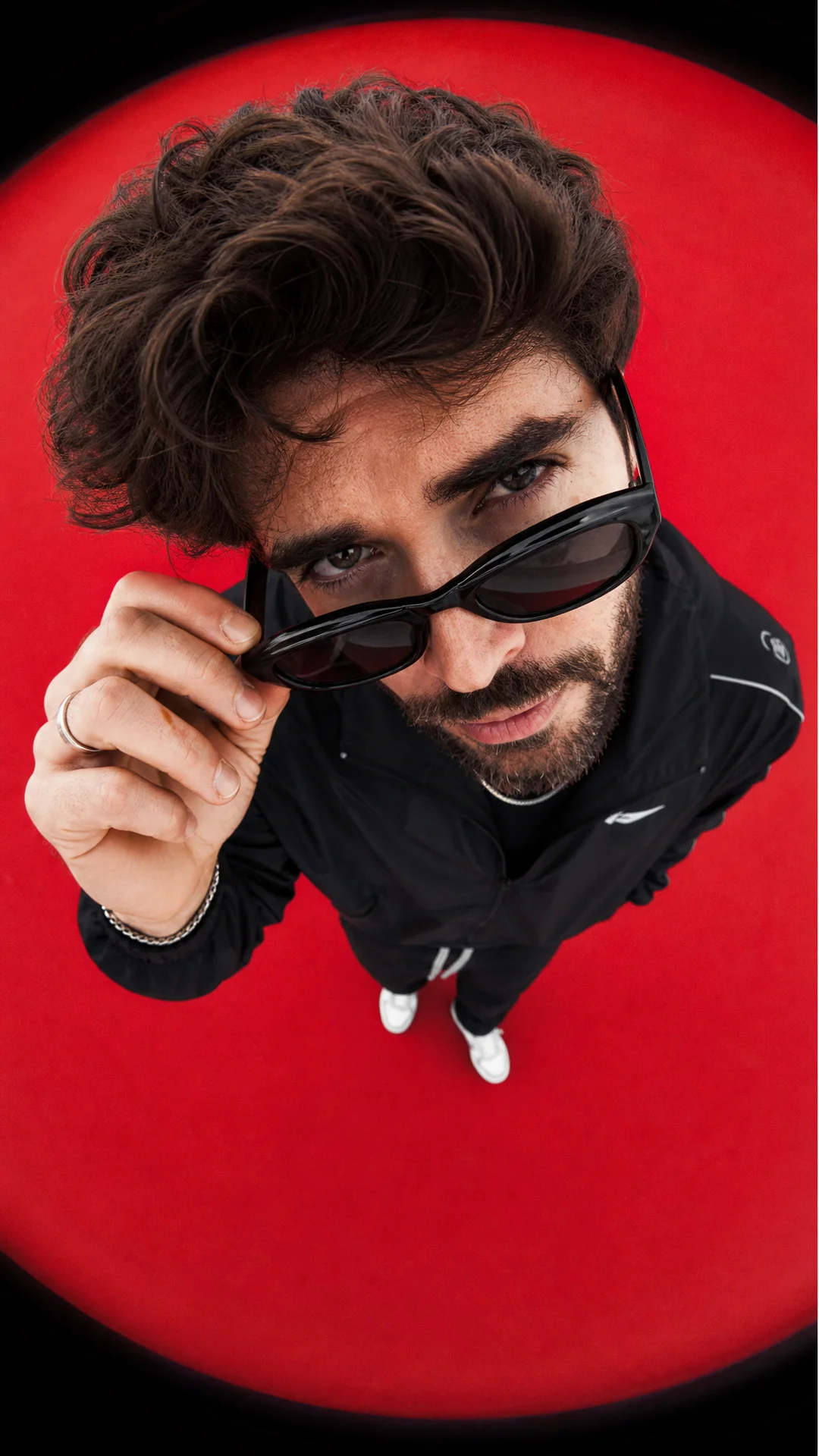 Экстремальная fashion-фотография с эффектом fisheye lens. Молодой мужчина с тёмными кудрявыми волосами стоит под камерой, камера расположена высоко над ним и направлена вертикально вниз. Он слегка наклоняется к объективу, лицо очень близко к камере, глаза