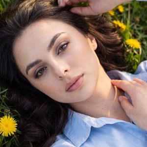 POV hyperrealistic 9:16 selfie, tight close-up while lying on a green meadow with yellow dandelions; invisible camera at arm’s length slightly above eye level, angled down; same outfit: light powder-blue cotton button-up with a soft collar, natural fabric