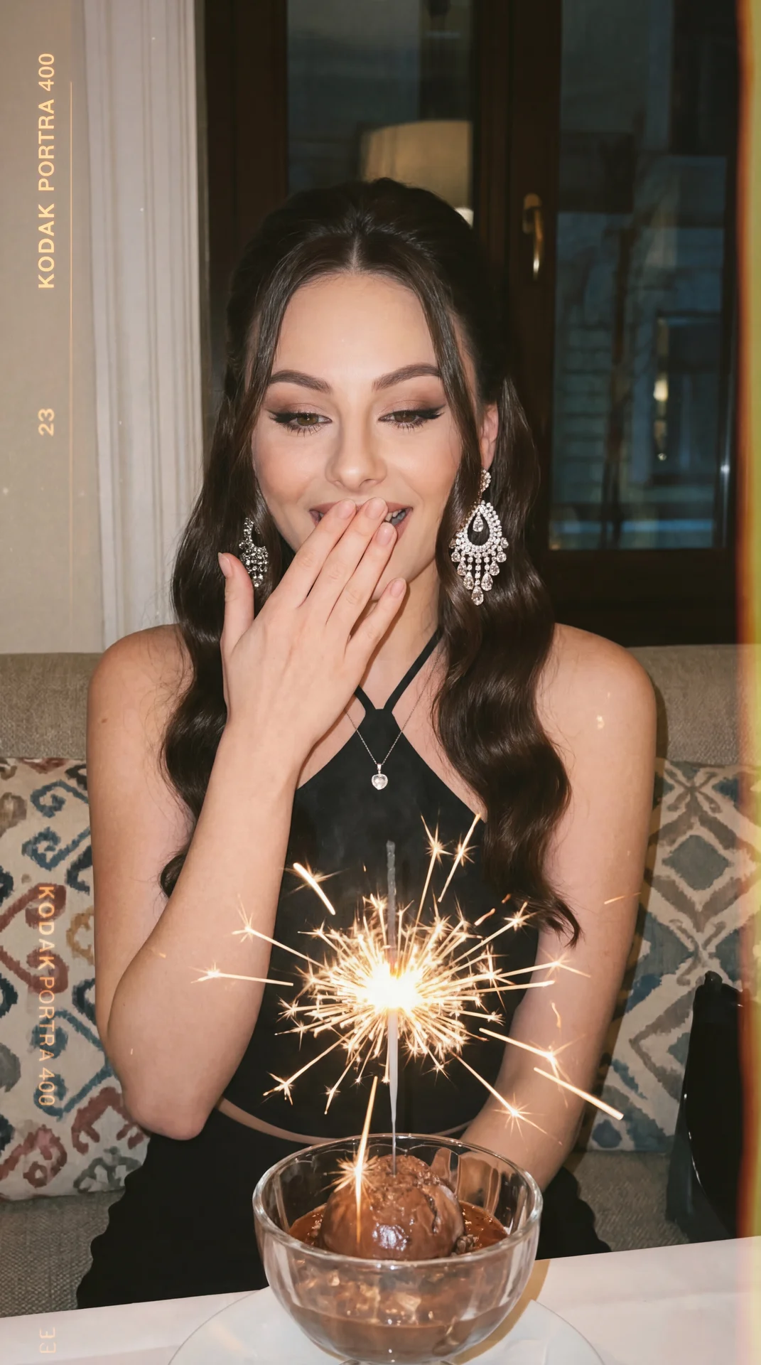 The image captures a joyful moment where a woman is reacting with delight. She is dressed elegantly in a black outfit and accessorized with sparkling, ornate earrings. Her long dark hair complements her look, and she wears a subtle necklace with a pendant