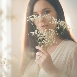 Крупный романтический портрет молодой женщины, лицо частично скрыто веточками гипсофилы с мелкими белыми цветами, холодные голубые глаза в центре, мягкий естественный макияж, нежные губы, тонкая рука с кольцом держит букет, сильно размытый передний план и