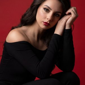 Professional studio portrait, tight three-quarter shot at eye level on a full-frame 85mm, woman in a matte black off-shoulder long-sleeve top and black pants, crouched with one knee raised and torso slightly forward; right forearm vertical resting on the 