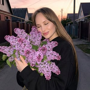 Максимально реалистичная фотография, лиричная вечерняя съемка на улице, ностальгическое и умиротворенное настроение.

Средний план. Девушка стоит на темной улице, прижимая к себе огромный букет пышной сирени. Глаза закрыты, голова слегка склонена к цветам