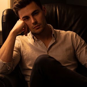 The image depicts a young man seated in an armchair, enveloped in a moody, dimly lit atmosphere. The lighting cast a warm, amber glow primarily from the left side, highlighting his features and casting deep shadows across the scene. He is wearing a light-