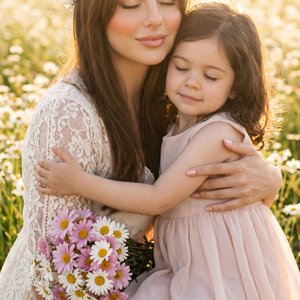 Young woman seated in a wildflower meadow at golden hour, gently embracing a little girl; woman’s left forearm cradles the child’s back and shoulder, right hand holding a bouquet of pink-and-white daisies near her waist; the child’s arms wrap around the w