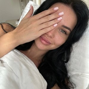 2.Эстетичное селфи молодой девушки с длинными волнистыми  волосами, лежащей в постели под белоснежным одеялом. Она кокетливо прикрывает лицо рукой с нежно-розовым маникюром квадрат, оставляя видимым один выразительный глаз и легкую улыбку. Ракурс сверху, 