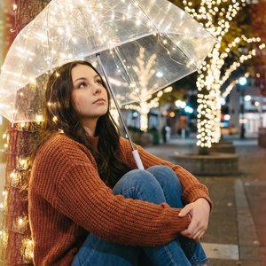 A girl is sitting on the ground outdoors at night, leaning gently against a tree wrapped with warm white fairy lights. She is holding a transparent umbrella that is also adorned with small, glowing fairy lights, creating a soft, magical illumination aroun