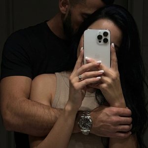 Интимное парное селфи в зеркале (mirror selfie), выполненное в стиле "low-key" с глубокими тенями и приглушенным освещением. Композиция крупного плана (medium close-up), фокусирующаяся на переплетении тел и рук. Ракурс прямой, имитирующий случайный домашн