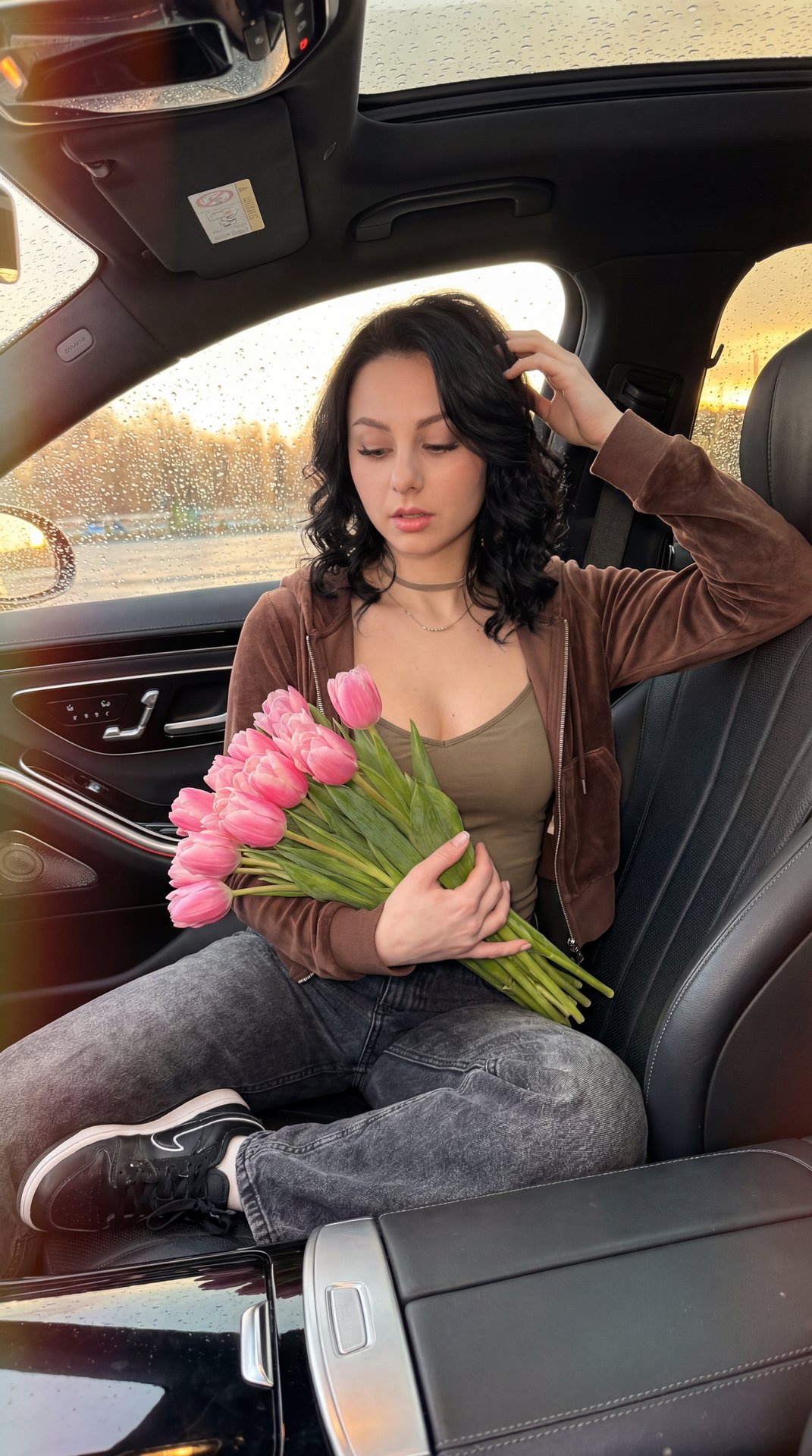 Young woman seated in the front passenger seat of a modern luxury car, medium shot from the driver’s side at a slightly elevated three‑quarter angle, professional camera look (full‑frame, 35mm, f/1.8, ISO 400, 1/200s), shallow depth of field, golden hour 