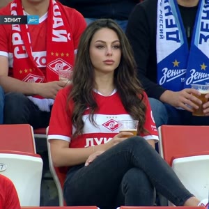 A realistic sports broadcast-style photo shoot taken in the stands of a stadium during a professional football match in Russia. The attached image shows a woman sitting in the stands. She has delicate features and a surprised yet focused expression, looki