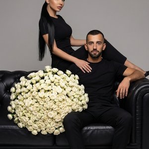 Не менять лица с фото. Стильный студийный портрет мужчины и женщины, сидящих на чёрном кожаном диване. Женщина сидит позади мужчины, её нога перекинута через его плечо, ладонь лежит у него на груди. Она в чёрном облегающем топе и брюках, с длинным прямым 