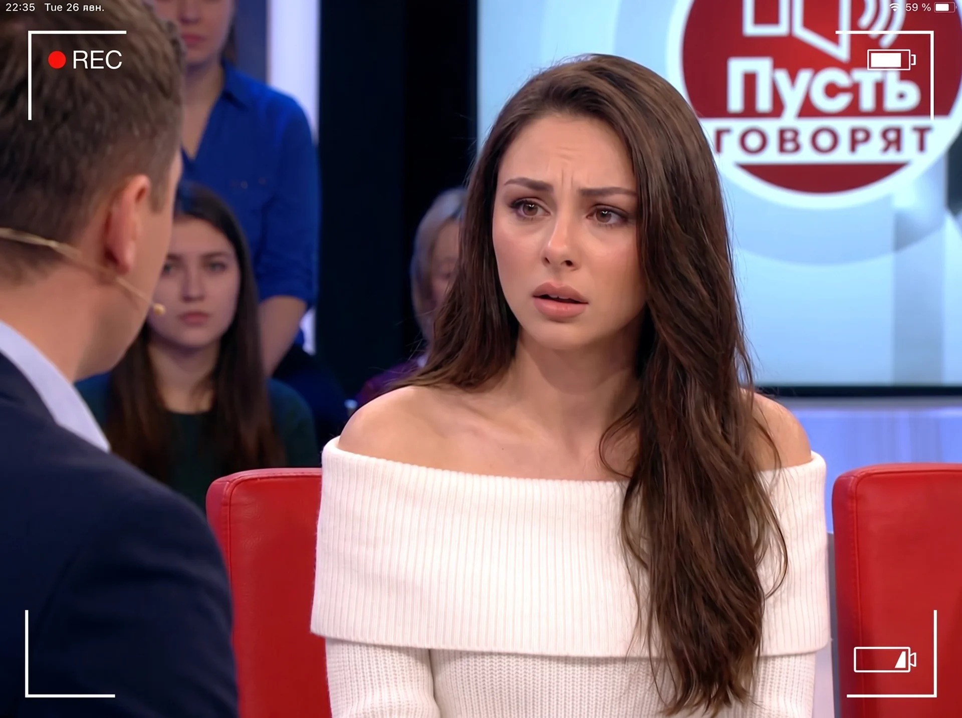 Young woman sitting in the guest chair on a dramatic Russian television talk show similar to “Пусть говорят,” filmed by a live studio broadcast camera. She is seated under bright studio lights, looking emotional and slightly surprised, with a serious expr
