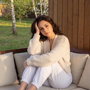 Hyperrealistic photography, indistinguishable from a real photo, raw photorealistic aesthetic. Female model seated curled up on an outdoor corner sofa on a wooden cabin porch in spring; camera at eye level, three-quarter medium shot from a slight front-ri