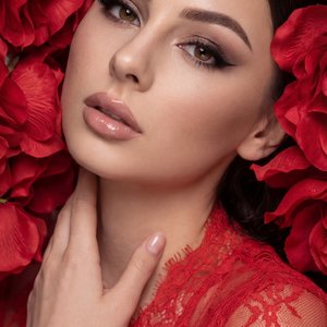 A girl is surrounded by vibrant red flower petals that frame her face closely, creating an immersive and dramatic floral backdrop. She is positioned leaning slightly backward, with only her upper body visible in the frame. Her right hand is gently touchin
