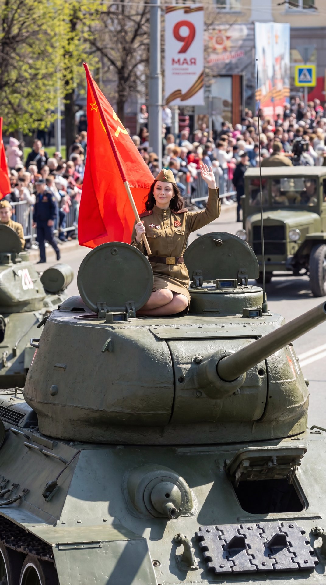 Hyperrealistic wide shot, a beautiful young woman in a WW2 Soviet military uniform sitting gracefully on the green armor of a T-34 tank. She is holding a red flag and waving. Sunny spring day, May 9th celebration, ultra-detailed metal textures, cinematic 