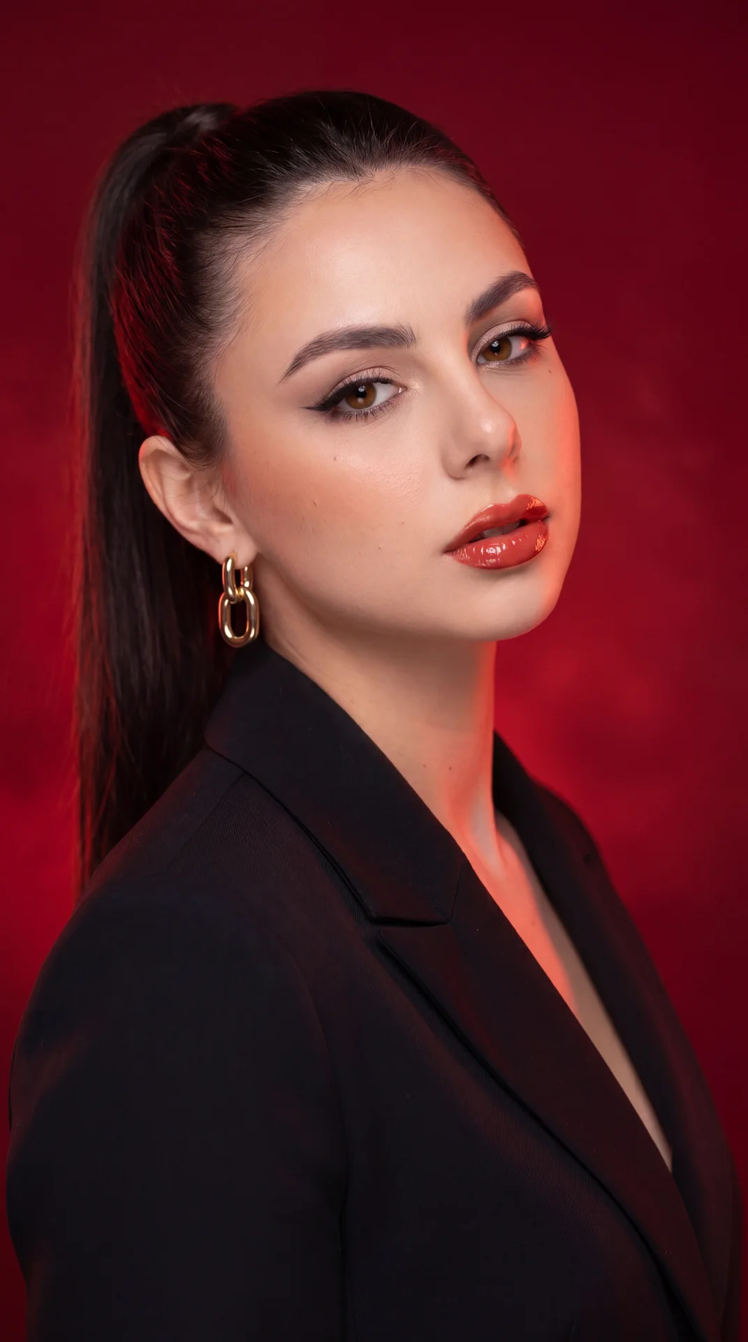 Professional studio portrait, medium close-up, three-quarter view; camera at eye level with a slight rightward offset; subject in a tailored matte black blazer (structured lapels), single chunky gold oval-link earring; hair in a sleek high ponytail. Torso