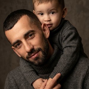 Не меняя внешности и волосы людей с фото, изображён мужчина и ребенок, связанные крепкой эмоциональной связью\. Мужчина смотрит в камеру, чуть наклонив корпус вперед\. Ребенок обнимает мужчину сзади за шею, смотрит с любопытством\. Мужчина держит рукой об