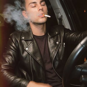 A men sitting in the driver’s seat of a car at night, medium close-up, shot from inside the cabin at a slight low angle with the steering wheel in the foreground framing the right side; head slightly tilted back, eyes gently closed, mouth slightly open ho