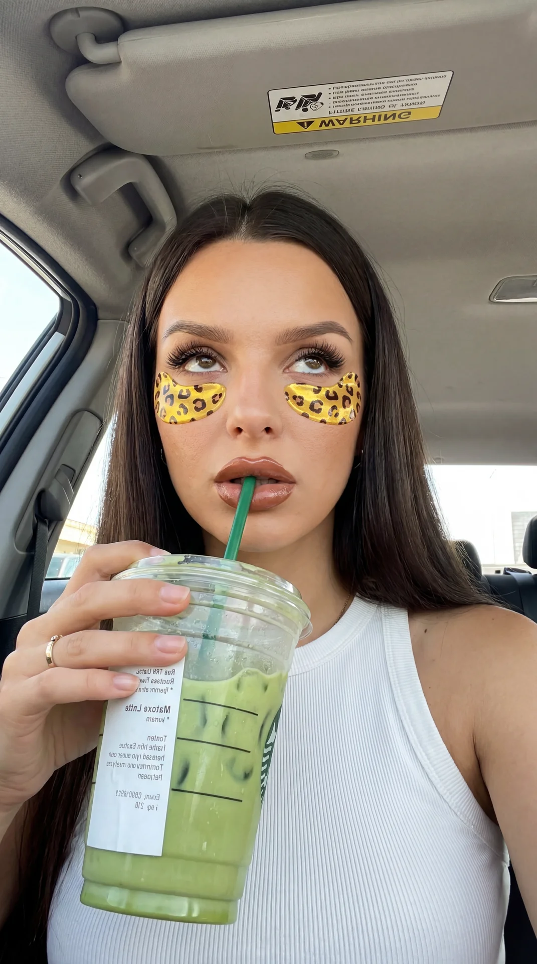A young woman sits in the passenger seat of a car, taking a selfie from slightly above and in front of her face, looking upward into the lens with a bored, slightly dramatic expression — eyes rolled slightly upward, full lips wrapped around a green reusab