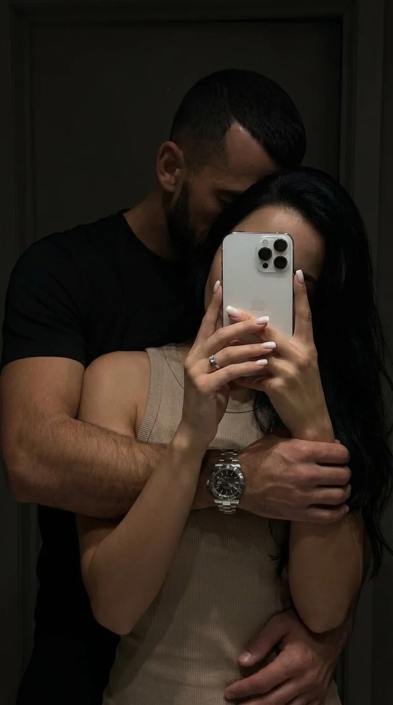 Интимное парное селфи в зеркале (mirror selfie), выполненное в стиле "low-key" с глубокими тенями и приглушенным освещением. Композиция крупного плана (medium close-up), фокусирующаяся на переплетении тел и рук. Ракурс прямой, имитирующий случайный домашн