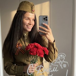 Девушка делает фото в зеркало на телефон. Она одета в советскую военную форму и на голове советская военная пилотка, на груди приколота Георгиевская ленточка. Одной рукой она держит букет красных гвоздик Фон нейтральный, стены серые, создавая спокойную ат