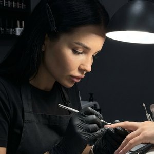 4.Create an image of a female nail master wearing a black professional uniform, sitting at her workstation in a bright nail studio. The room is mostly dark except for a focused lamp light shining over the workspace. Her hair is pinned to one side with a b