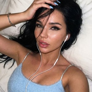 Не меняй черты лица.
Молодая женщина лежит на белой подушке в расслабленной позе. Густые волосы рассыпаны по подушке, несколько прядей падают на лицо. Она игриво смотрит прямо в камеру, одна рука в волосах.
Макияж: чёрные графичные стрелки «cat eye», длин