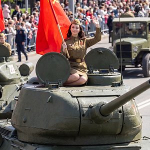 Hyperrealistic wide shot, a beautiful young woman in a WW2 Soviet military uniform sitting gracefully on the green armor of a T-34 tank. She is holding a red flag and waving. Sunny spring day, May 9th celebration, ultra-detailed metal textures, cinematic 