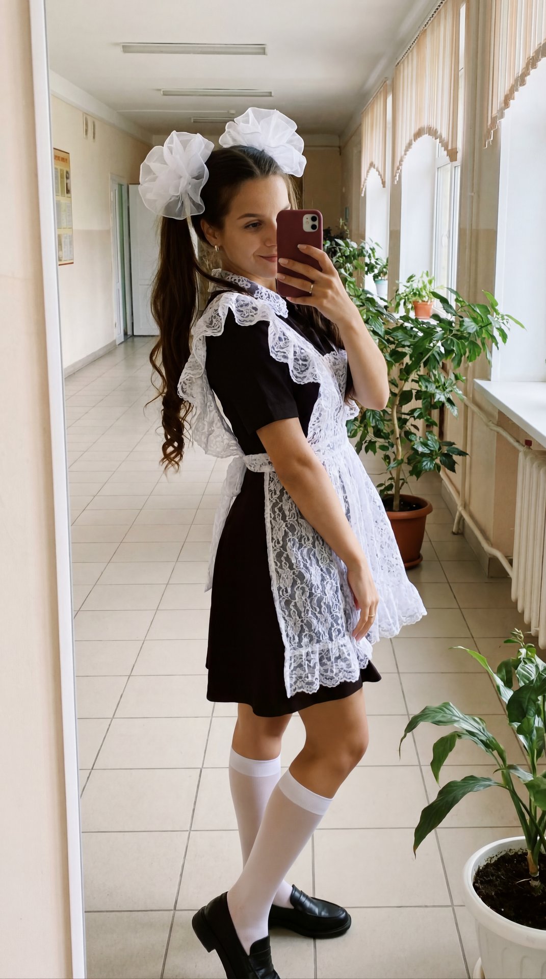 Создай Максимально реалистичное mirror selfie фото в школьном коридоре, одна девушка-выпускница стоит напротив большого зеркала и фотографирует себя на телефон. На ней советская школьная форма: приталенное чёрное платье с короткими рукавами и белый очень 