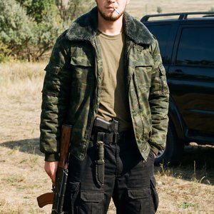 Высококачественное фото молодого мужчины в стиле military. На нем объемная темно-зеленая камуфлированная куртка-бомбер с воротником, надетая поверх оливковой базовой футболки, и широкие черные тактические штаны с карманами, на правом бедре тактический нож