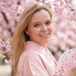 Ultra-detailed portrait of a young woman with a cute and charming smile, looking towards the camera. She is wearing a soft pastel pink traditional kimono, standing under blooming cherry blossom trees (sakura), cinematic spring atmosphere. She is gently tu