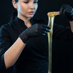 1,2.Create an image of a female depilation master in a dark studio with a black background. She wears professional black attire and black gloves. In her hand, she holds a wooden spatula, stretching a thick strand of warm golden wax from a white container.