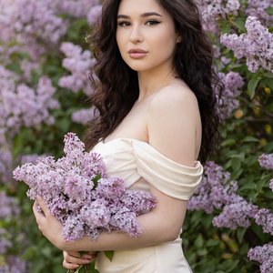 A woman with long softly curled hair stands slightly sideways to the camera among blooming lilac bushes. She is not wearing a hat, allowing her hair to flow naturally around her shoulders.

Shot Type:
Shot from the waist up, the woman is positioned slight