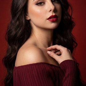 Studio portrait, elegant feminine silhouette, three‑quarter pose with shoulders angled, head slightly tilted; gaze directed to camera‑left; lips softly parted with a subtle confident expression. Right hand raised to the collarbone, fingertips lightly touc