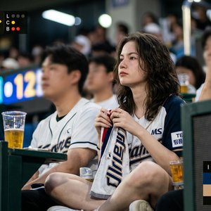 Use the attached photo as the exact facial reference. Create an ultra realistic 16:9 Korean baseball broadcast crowd shot during a summer evening {팀 이름} game. Preserve the original face and hairstyle naturally without over-editing. Wide shot from an eye-l