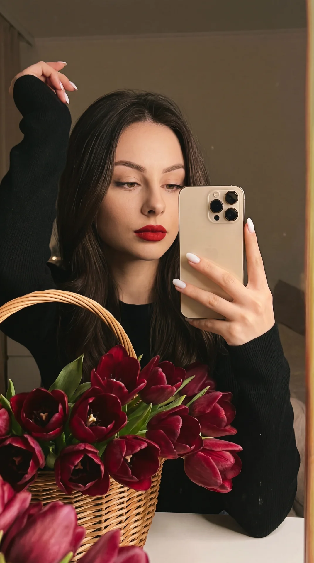 А girl taking a mirror selfie indoors, medium close-up shot with a slight high angle and three-quarter framing; right hand raised and bent at the elbow, holding a metallic gold smartphone with a triple-lens module near cheek level, fingers wrapped around 