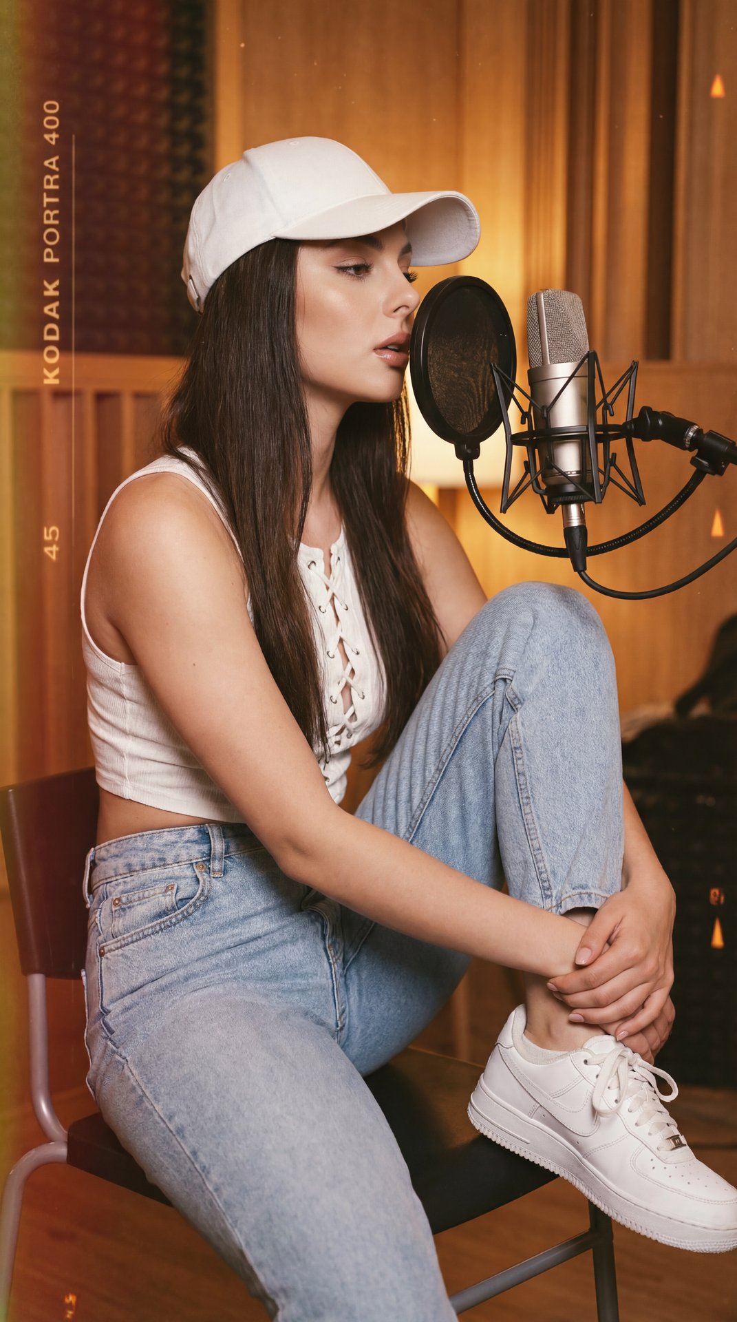 A girl is seated on a black chair in a cozy, wood-paneled recording studio, captured in a medium close-up shot. The camera angle is slightly from the side, eye-level, focusing on her upper body and face as she leans casually forward toward a recording mic