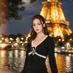 А girl, three-quarter shot, slightly low angle, professional DSLR look (35mm, wide aperture, crisp subject, soft bokeh), night by the Seine on a stone embankment, Eiffel Tower glowing warm gold behind, full moon through tree branches, reflections on water
