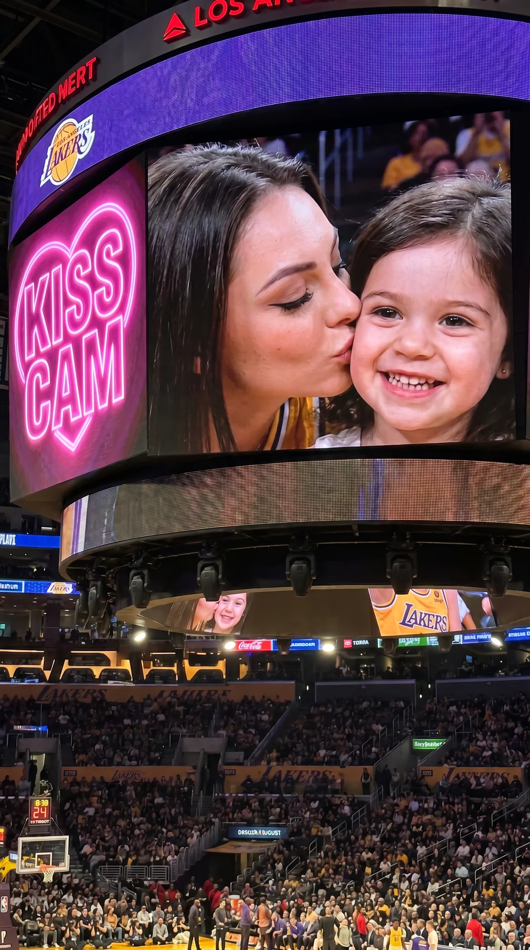 Young woman sitting at a Lakers playoff game shown on the giant arena screen during a kiss cam moment. Camera is closer to the jumbotron so her face is clearly visible and fills most of the frame. She is leaning in and affectionately kissing a cute young 