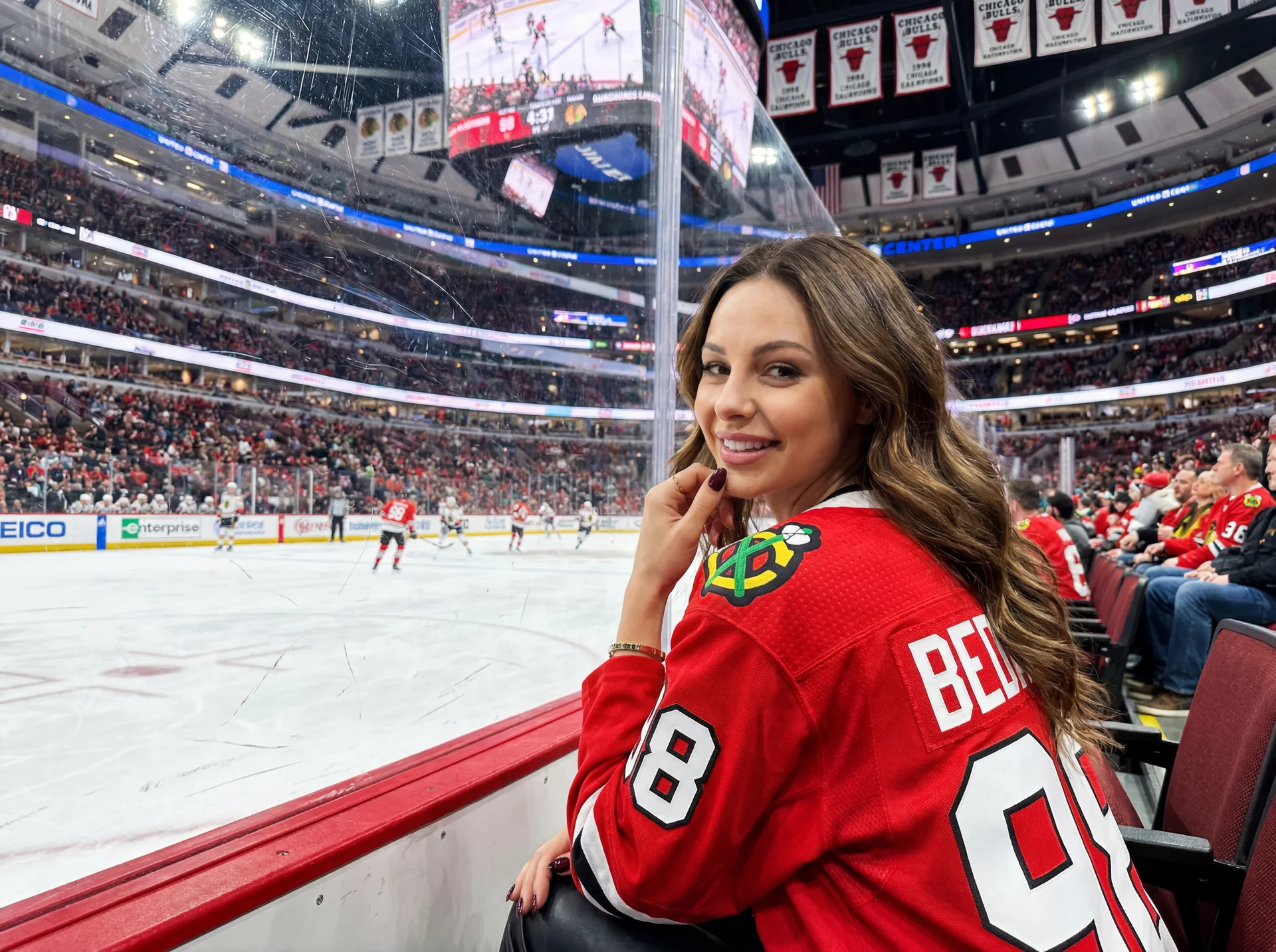 Create a realistic high quality photo using the face from the reference without any changes or distortions. Realistic lifestyle photo of a young woman at a hockey game in a huge modern arena. The atmosphere is lively energetic and flirty with American spo