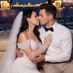 Newlywed couple on a yacht at night, medium close-up, eye-level, straight-on, professional editorial wedding photo, full-frame camera, 35mm prime, f/1.8, shallow depth of field, warm diffused flash fill from front-left; woman on the right in a white off-s
