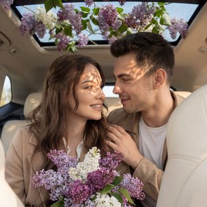 Фотореалистичная сцена в салоне автомобиля с панорамным люком, через который свисают ветки сирени разных оттенков (белая, светло-фиолетовая, тёмно-фиолетовая). Молодая пара сидит на заднем сиденье в уютных бежевых пальто — они смотрят друг другу в глаза, 