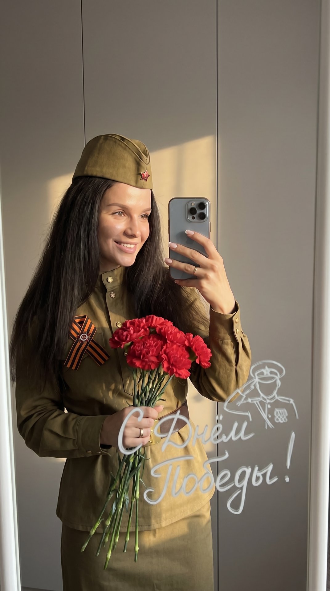 Девушка делает фото в зеркало на телефон. Она одета в советскую военную форму и на голове советская военная пилотка, на груди приколота Георгиевская ленточка. Одной рукой она держит букет красных гвоздик Фон нейтральный, стены серые, создавая спокойную ат