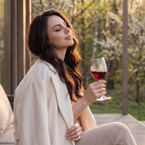Hyper-realistic authentic raw photograph, completely indistinguishable from reality, unretouched lifestyle capture. Young woman seated sideways on an outdoor lounge at a modern wood-and-glass cabin patio, fresh and beautiful spring afternoon. Medium three
