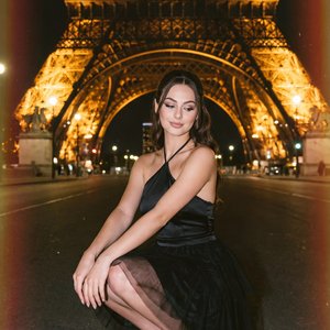 А girl crouching centered under the illuminated Eiffel Tower at night, medium-full shot (full figure while crouched) from a slightly low, frontal angle with symmetrical framing; she balances on black strappy stiletto sandals, knees together, torso upright